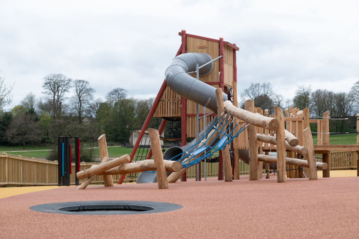 Wooden playground with climbing frames, rope bridge, and tube slide on rubber surface.